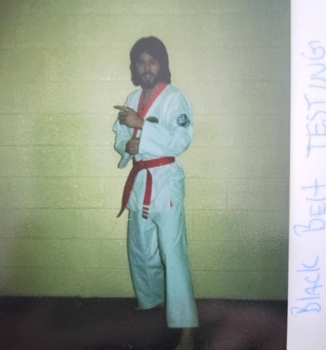 A young Michael Birch with long hair and a beard, in a white uniform and red belt, pointing toward the camera. "Black Belt Testing" is handwritten in the margin of the print.