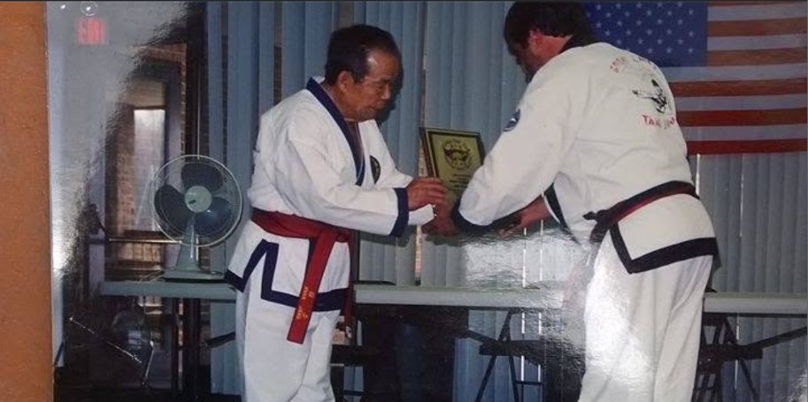Master Birch, in a black-trimmed uniform, hands a plaque to Grandmaster Jae Joon Kim in front of an American flag.